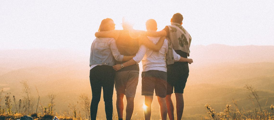 Groups of 4 friends with their arms around each other's shoulders and backs looking into the sunset.